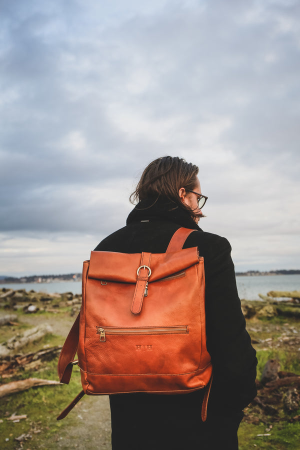 Reinald Roll Top leather backpack in Cognac. Worn by male model in black coat at beach with driftwood and grass and Pacific ocean. 