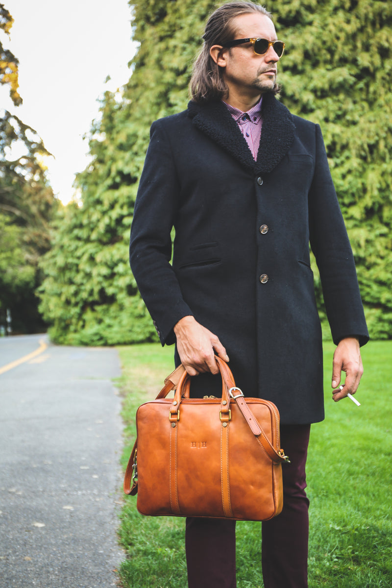 Piero Briefcase in Cognac. Held by male model with long brown hair, sunglasses, and holding a cigarette. Handcrafted Italian leather. 